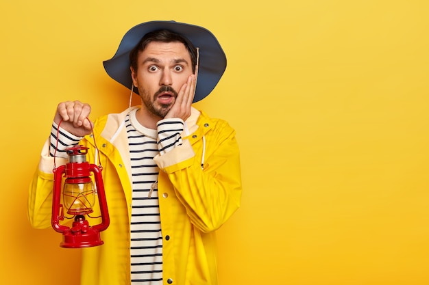 Scared male traveler holds little red lantern for lightening in darkness, keeps palm on cheek, wears waterproof yellow raincoat