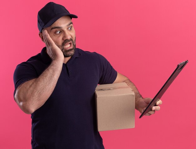 Scared middle-aged delivery man in uniform and cap holding box looking at clipboard in his hand isolated on pink wall