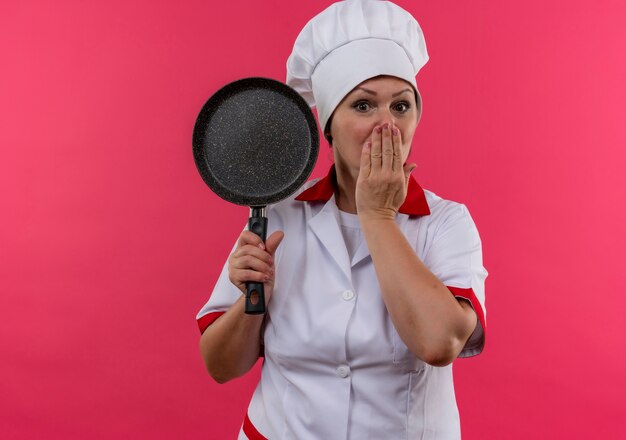 Scared middle-aged female cook in chef uniform holding frying pan covered mouth with hand with copy space