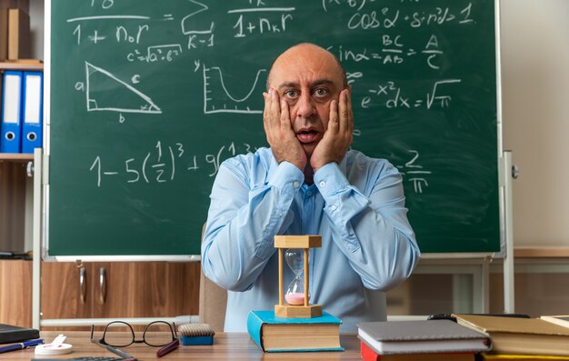 scared middle-aged male teacher sits at table with school supplies covered face with hands in classroom
