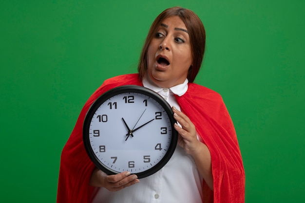 Scared middle-aged superhero female looking at side holding wall clock isolated on green