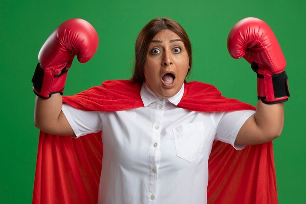 Scared middle-aged superhero female wearing boxing gloves raising hands isolated on green