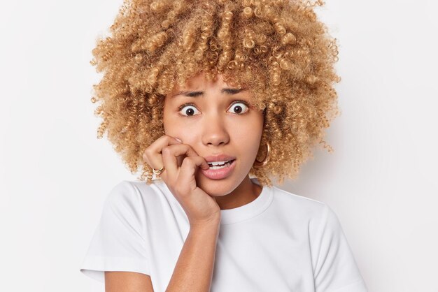 Scared nervous woma bites fingernails feels anxious embarrassed about something dressed in casual t shirt poses against white background being troubled stands insecure. Bad feelings concept.