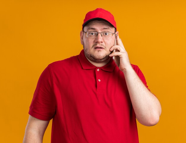 scared overweight young deliveryman in optical glasses talking on phone isolated on orange wall with copy space