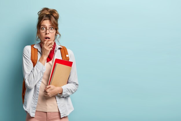 Scared overwhelmed woman gazes surprisingly at camera, has bated breath, holds necessary notepads