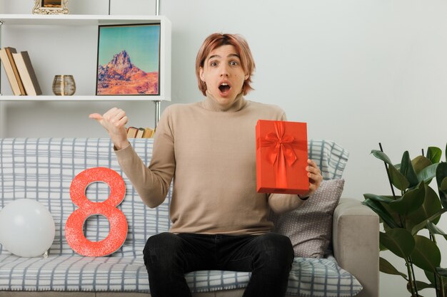 Scared points at side handsome guy on happy women day holding present sitting on sofa in living room