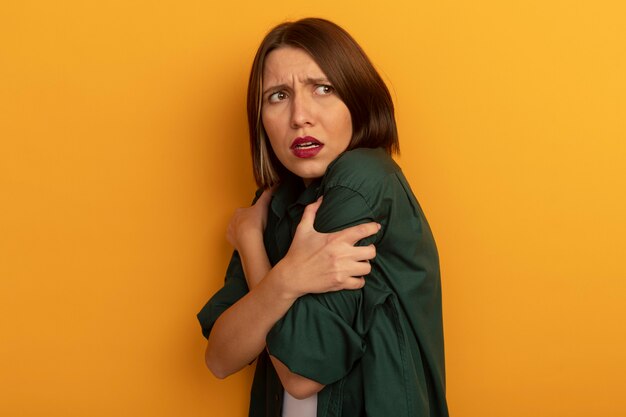 Scared pretty caucasian woman stands sideways holding arms and looking at side on orange