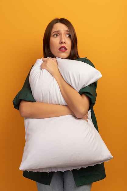 Free photo scared pretty woman holds pillow and looks at side isolated on orange wall