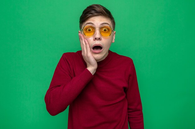 Free photo scared putting hand on cheek young handsome guy wearing glasses isolated on green wall