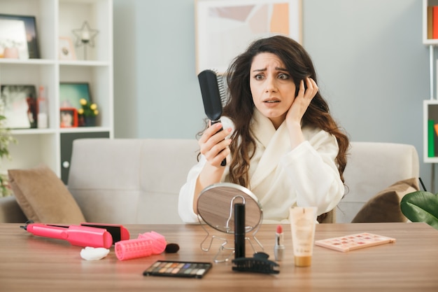 Scared putting hand on head young girl holding and looking at comb sitting at table with makeup tools in living room