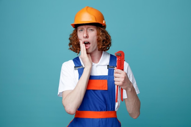 Испуганный кладущий руку на рот молодой строитель в форме, держащий газовый ключ на синем фоне