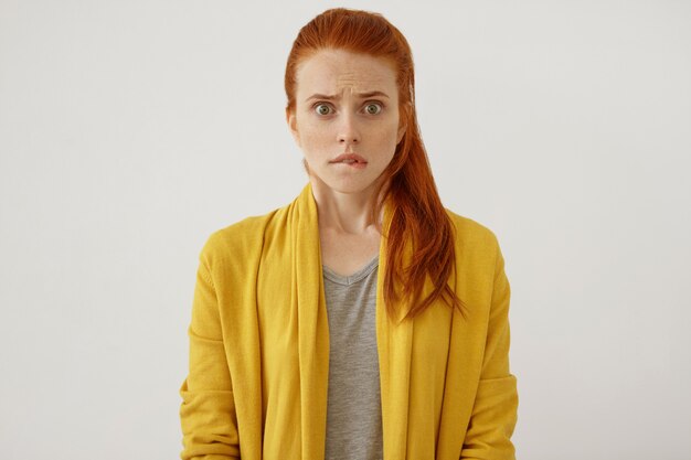Scared red-haired woman with pony tail, stairing, curving her lips being shocked to realize something horrorful. Shocked young female in yellow clothes isolated