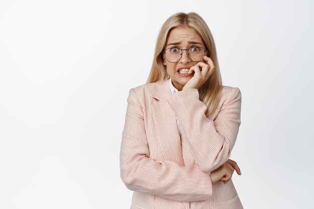 Scared saleswoman biting fingernatils looking frightened at camera anxious of something standing in suit and glasses over white background