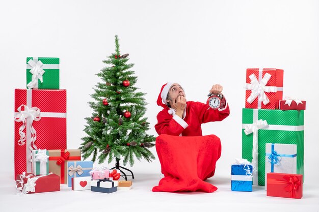 Scared santa claus in deep thoughts sitting on the ground and showing clock near gifts and decorated xsmas tree on white background