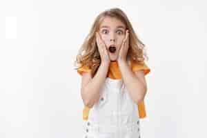 Free photo scared and shocked caucasian blond little girl gasping, grab face amazed, stare camera astonished, drop jaw speechless observe shook event, react ambushed, frightened see spider, white wall