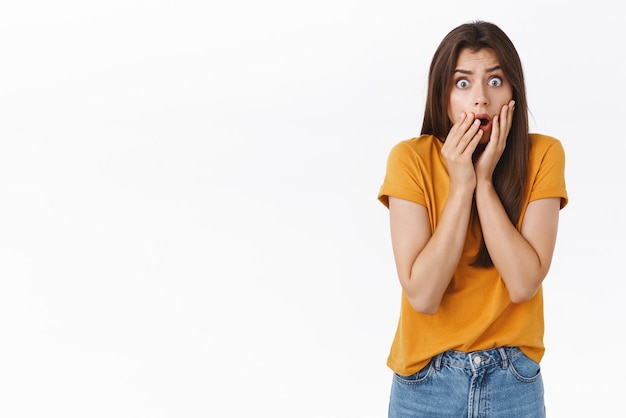 Scared shocked timid insecure woman standing speechless drop jaw gasping stare camera frightened hear stunning rumours holding hands on mouth horrified standing white background