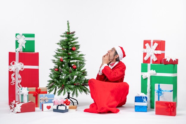 Испуганный шокированный молодой человек, одетый как Санта-Клаус с подарками и украшенной елкой, сидит на земле на белом фоне