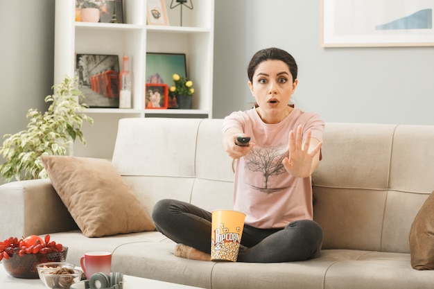 Scared showing stop gesture young girl holding tv remote, sitting on sofa behind coffee table in living room