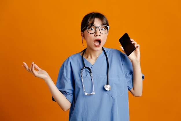 Испуганная раскинутая рука держит телефон молодой женщины-врача в униформе, изолированной на оранжевом фоне