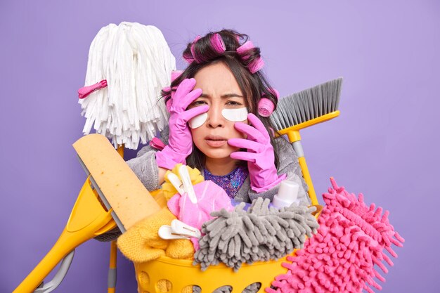 Scared stressed Asian woman housekeeper applies collagen pads under eyes has frightened expression keeps hands on face makes hairstyle poses near laundry basket isolated over purple background.