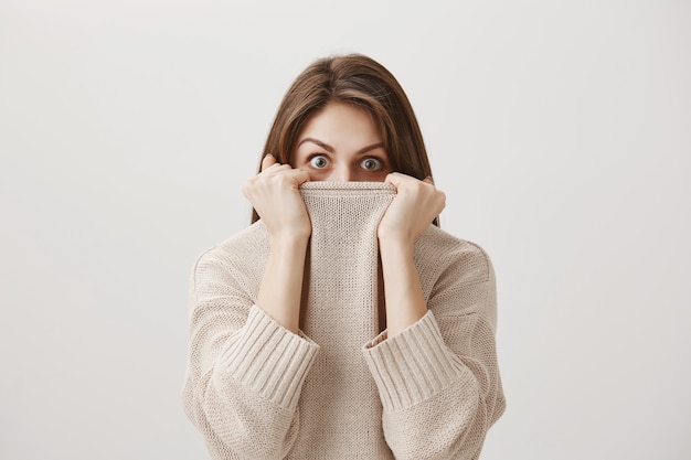 Scared timid girl hiding face in sweater collar and alarmed