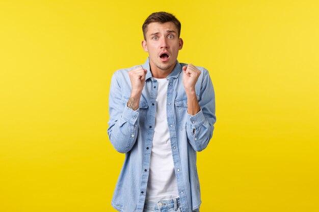 Scared timid guy trembling from fear, looking worried and nervous, shivering and staring concerned camera, raising fists up clumsy. Weak man trying defend himself from assault, yellow background