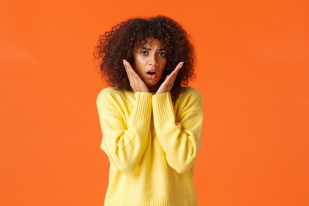 Scared timid and insecure lovely young girl with afro haircut, yellow sweater, gasping shocked and concerned, touching and staring in panic , feeling afraid, standing orange wall.