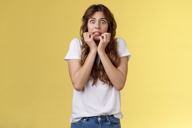 Scared timid insecure unconfident european girl curly haircut hold hands face biting fingers look frightened shocked standing stupor speechless shaking fear terrified yellow background