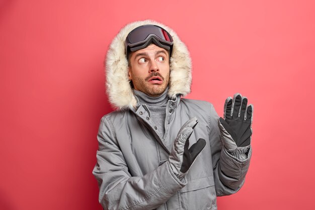 Scared winter man makes defensive gesture afraids as something heavy going to fall on him wears grey jacket with fur hood and ski goggles.