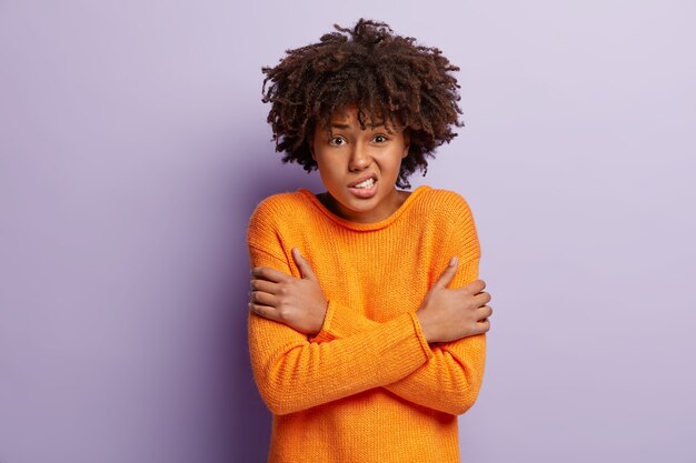scared woman trembles from fear, crosses hands over chest, feels freezing, clenches teeth, wears orange casual jumper, isolated over purple wall, frowns face. unhappy girl feels cold indoor