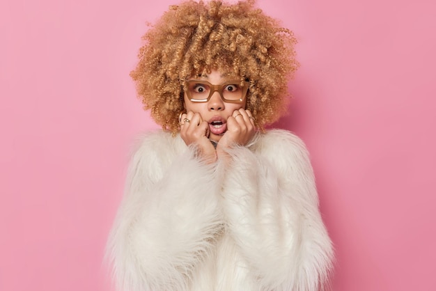 Scared worried curly haired woman keeps hand on face looks concerned and frightened stares shocked at camera wears spectacles white fur coat isolated over pink background. Human reactions concept