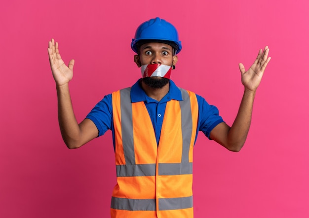 Scared young afro-american builder man in uniform with safety helmet mouth sealed with warning tape standing with raised hands isolated on pink background with copy space