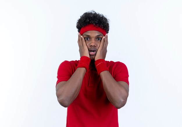 Scared young afro-american sporty man wearing headband and wristband putting hands on cheek isolated on white