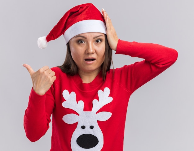 Scared young asian girl wearing christmas hat with sweater points at side putting hand on head isolated on white background