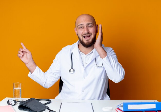 Scared young bald male doctor wearing medical robe and stethoscope sitting at work desk