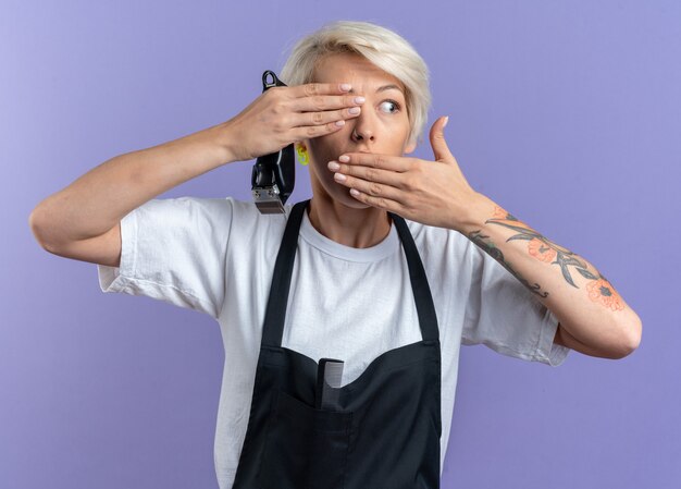 Scared young beautiful female barber in uniform holding hair clippers covered eye and mouth isolated on blue background