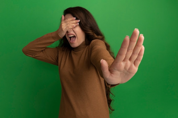 Free photo scared young beautiful girl covered eyes with hands and holding out hand  isolated on green wall