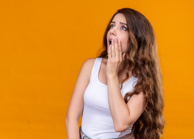 Scared young beautiful girl looking at side with hand near mouth on isolated orange space with copy space