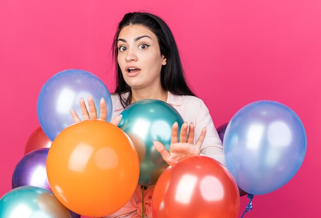 Scared young beautiful girl standing behind balloons holding hands at camera 
