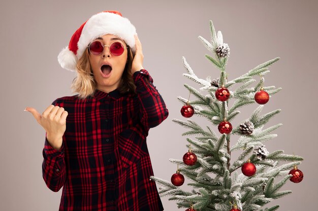 Scared young beautiful girl standing nearby christmas tree wearing christmas hat with glasses points at side isolated on white background with copy space