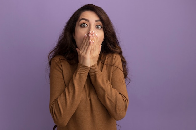 Scared young beautiful girl wearing brown turtleneck sweater covered face with hands isolated on purple wall