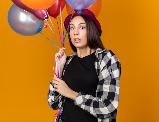 Scared young beautiful girl wearing party hat holding balloons isolated on orange wall