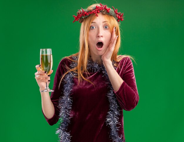 Scared young beautiful girl wearing red dress with wreath and garland on neck holding glass of champagne putting hand on cheek isolated on green background