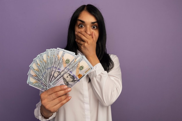 Free photo scared young beautiful girl wearing white t-shirt holding money covered mouth with hand isolated on purple with copy space