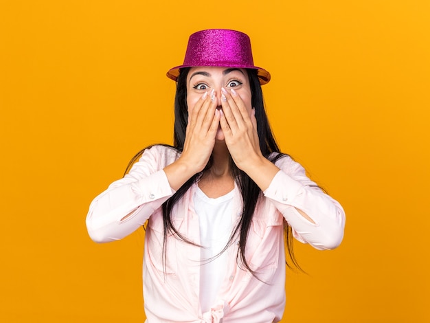 Scared young beautiful woman wearing party hat covered face with hands isolated on orange wall