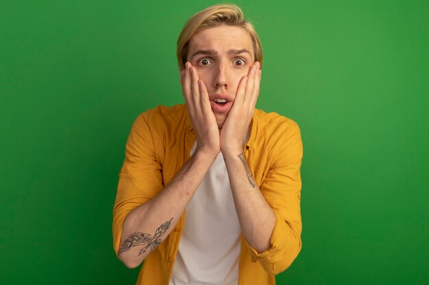 Scared young blonde guy wearing yellow t-shirt covered face with hands isolated on green