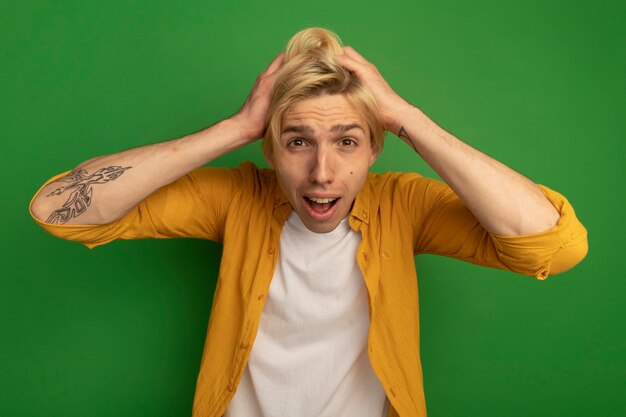 Scared young blonde guy wearing yellow t-shirt grabbed head isolated on green