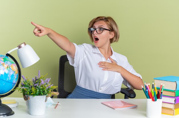 Scared young blonde student girl wearing glasses sitting at desk with school tools keeping hand on chest looking and pointing at side isolated on olive green wall