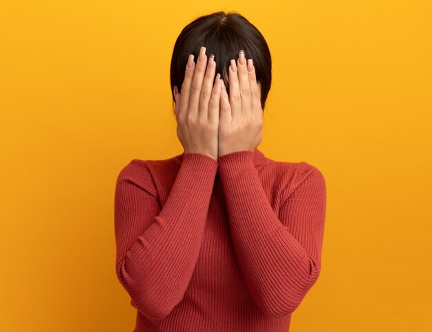 Scared young brunette caucasian girl covers face with hands isolated on orange wall with copy space