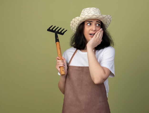 Scared young brunette female gardener in uniform wearing gardening hat puts hand on face and holds rake looking at side isolated on olive green wall